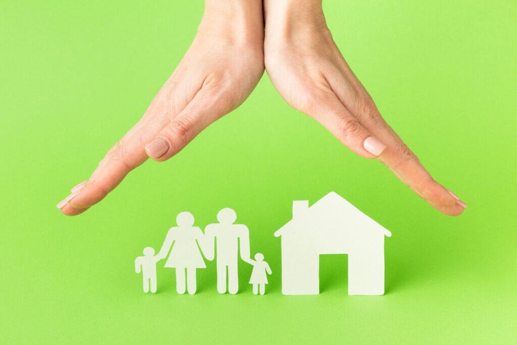Hands protecting paper cutouts of a family and house. Life insurance protection.