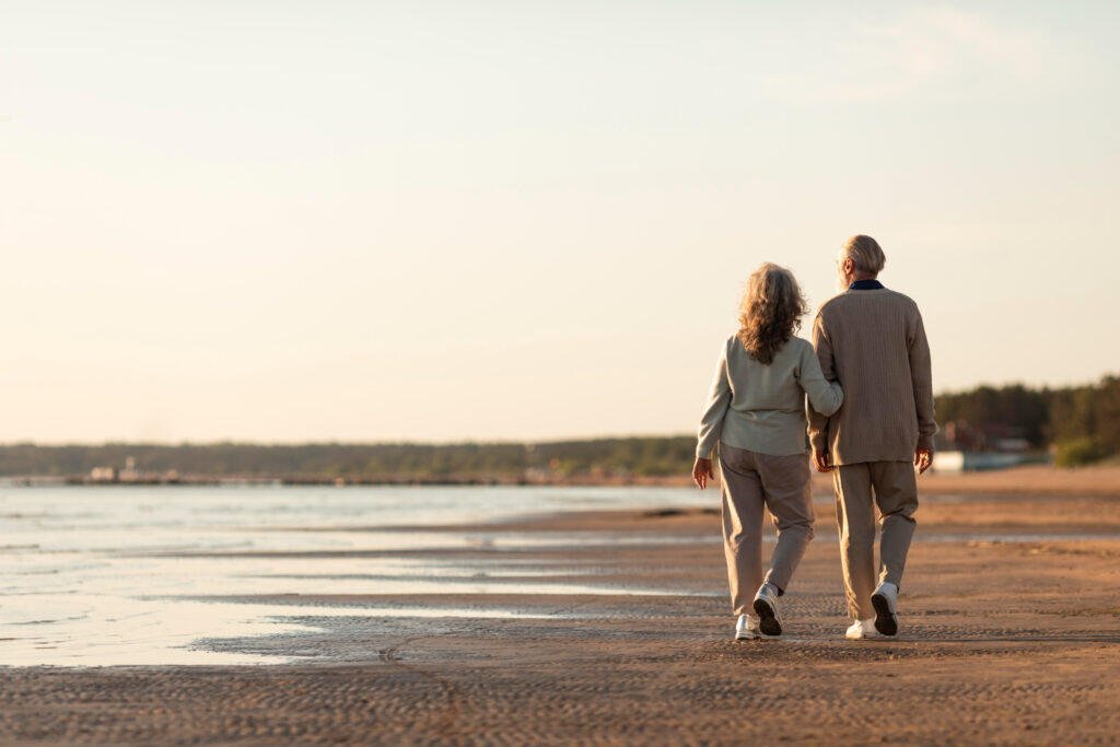 Happy retired couple holding hands, walking on a beach at sunset.