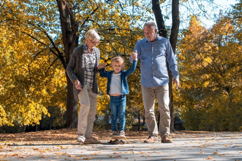 Grandparents helping granddaughter learn to skateboard in autumn park.
