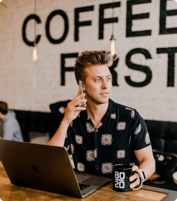 Man on phone call in coffee shop, working on laptop